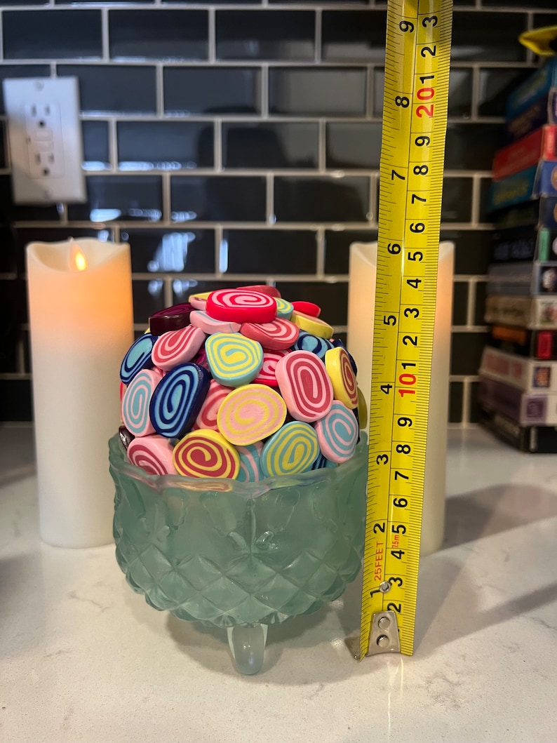 May include: A teal glass pedestal with three legs holds a cluster of colourful, swirl-shaped sweets. The sweets are in shades of pink, blue, yellow, and green. A yellow measuring tape is visible to the right of the pedestal.