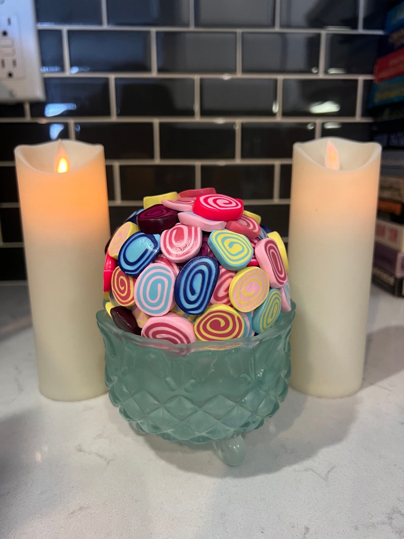 May include: A teal glass bowl with three legs holds a collection of colourful swirl-shaped sweets. The sweets are in shades of pink, blue, yellow, and red.