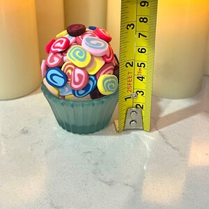 May include: A blue cupcake liner filled with colorful swirl candies. The candies are pink, yellow, blue, and brown. The cupcake liner is sitting on a white surface next to a yellow measuring tape.