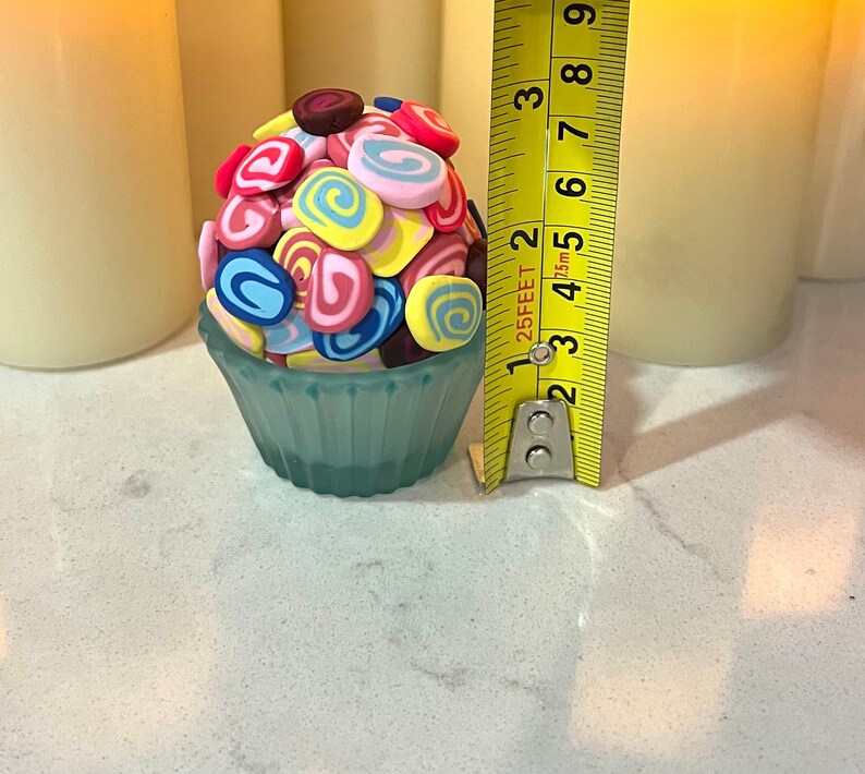 May include: A blue cupcake liner filled with colorful swirl candies. The candies are pink, yellow, blue, and brown. The cupcake liner is sitting on a white surface next to a yellow measuring tape.