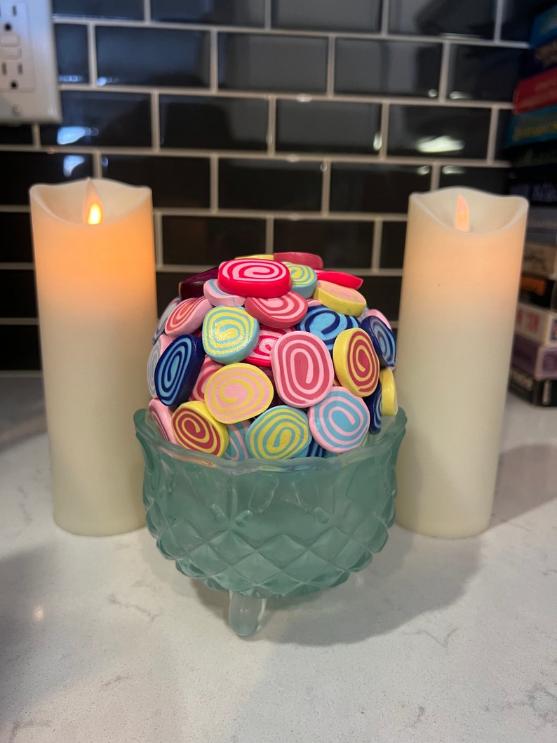 May include: A teal glass bowl with a decorative diamond pattern sits on a white surface. The bowl is filled with colourful, swirl-shaped sweets. Two white candles are on either side of the bowl.