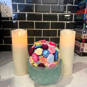 May include: A teal glass bowl with a decorative design sits on a white countertop. The bowl is filled with colourful, swirl-shaped sweets. Two white candles are on either side of the bowl.