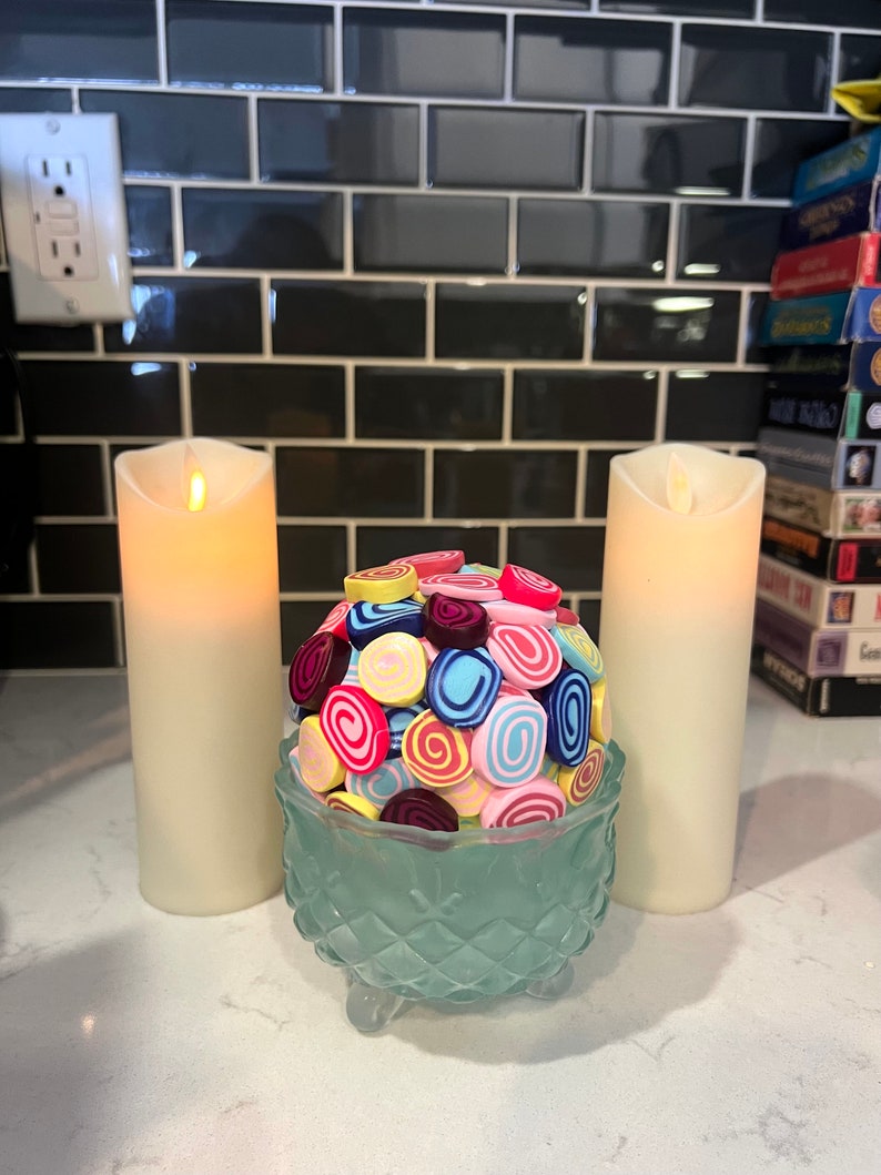 May include: A teal glass bowl with a decorative design sits on a white countertop. The bowl is filled with colourful, swirl-shaped sweets. Two white candles are on either side of the bowl.