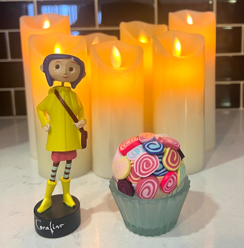 May include: A yellow raincoat figurine of Coraline Jones from the movie Coraline, standing on a black base with the word "Coraline" written on it.  The figurine is in front of a group of five white pillar candles.  A small cupcake with colorful swirl decorations is in the foreground.
