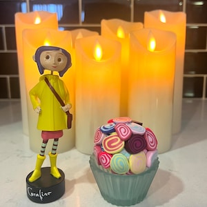 May include: A figurine of Coraline from the movie Coraline, wearing a yellow raincoat and holding a brown bag. The figurine is standing in front of five white pillar candles with a yellow glow. A cupcake with colorful frosting is in the foreground.