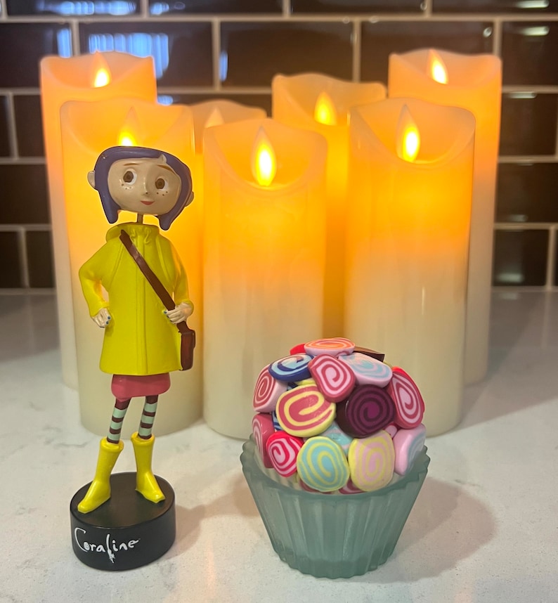 May include: A figurine of Coraline from the movie Coraline, wearing a yellow raincoat and holding a brown bag. The figurine is standing in front of five white pillar candles with a yellow glow. A cupcake with colorful frosting is in the foreground.