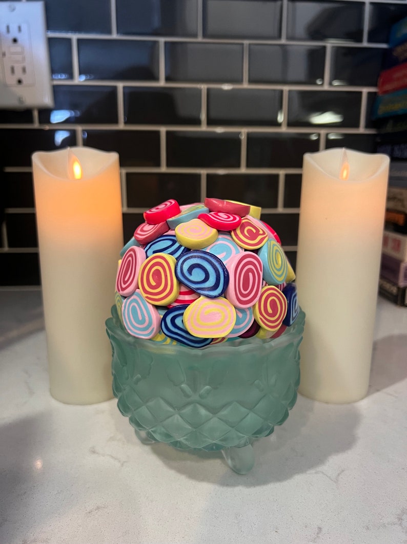 May include: A teal glass bowl with three legs sits on a white countertop. The bowl is filled with colourful, swirl-shaped sweets. Two white candles are on either side of the bowl.