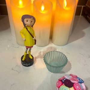 May include: A figurine of Coraline from the movie Coraline, wearing a yellow raincoat, standing in front of five lit candles. A small blue glass bowl and a ball of colorful candy are also in the image.