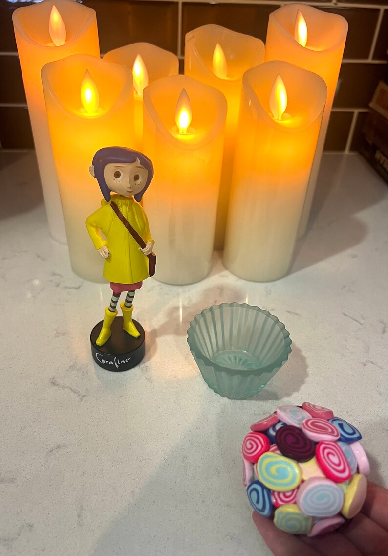 May include: A figurine of Coraline from the movie Coraline, wearing a yellow raincoat, standing in front of five lit candles. A small blue glass bowl and a ball of colorful candy are also in the image.
