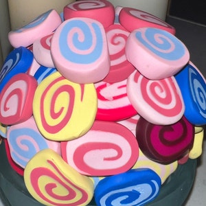 May include: A close-up of a colorful arrangement of swirl-shaped candies, featuring pink, blue, yellow, red, and white colors. The candies are arranged in a circular pattern, creating a visually appealing display.