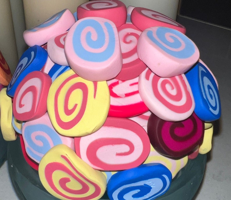 May include: A close-up of a colorful arrangement of swirl-shaped candies, featuring pink, blue, yellow, red, and white colors. The candies are arranged in a circular pattern, creating a visually appealing display.