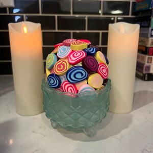 May include: A glass bowl with a decorative design sits on a white countertop. The bowl is filled with colourful, swirl-shaped sweets. Two white candles are on either side of the bowl.