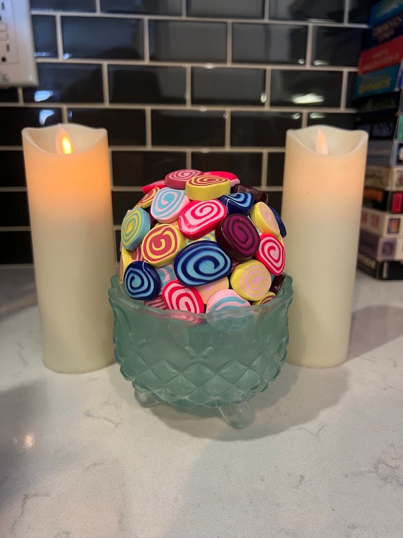 May include: A glass bowl with a decorative design sits on a white countertop. The bowl is filled with colourful, swirl-shaped sweets. Two white candles are on either side of the bowl.