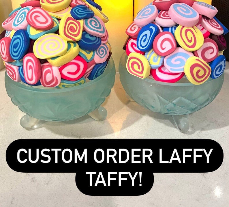 May include: Two glass bowls with three legs each, filled with colorful swirled Laffy Taffy candies. The bowls are on a white surface with the text "CUSTOM ORDER LAFFY TAFFY!" below them.