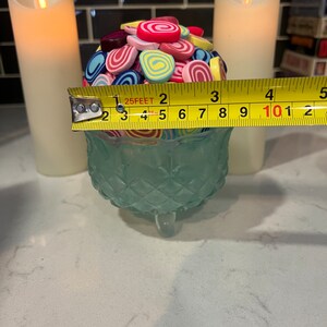 May include: A teal glass bowl with a decorative pattern sits on a white countertop. The bowl is filled with colourful, swirl-shaped sweets. A yellow measuring tape is placed across the top of the bowl, with the numbers 1, 2, 3, 4, and 5 visible. Two white candles are in the background.
