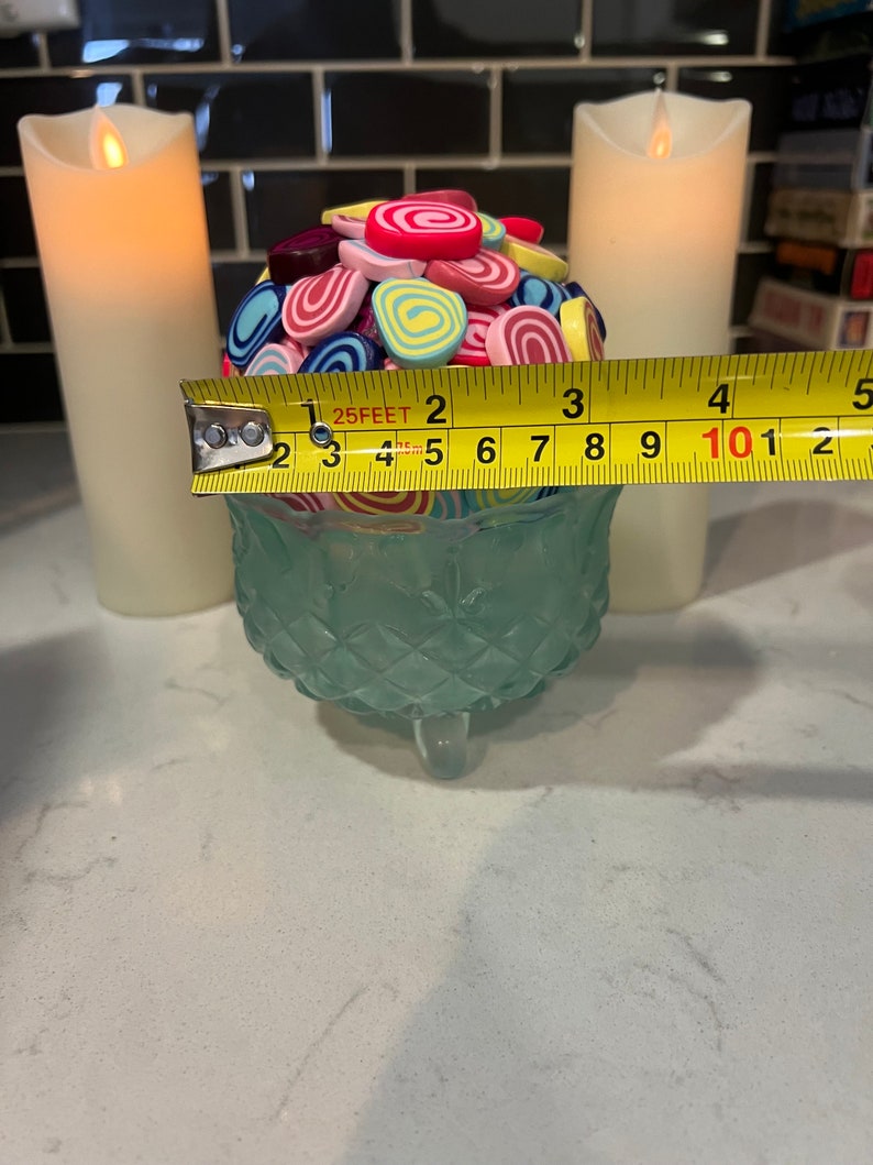 May include: A teal glass bowl with a decorative pattern sits on a white countertop. The bowl is filled with colourful, swirl-shaped sweets. A yellow measuring tape is placed across the top of the bowl, with the numbers 1, 2, 3, 4, and 5 visible. Two white candles are in the background.