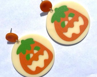 May include: A pair of circular earrings featuring a cream-colored base with a central orange jack-o'-lantern graphic. Each pumpkin design includes a green stem and a wavy-edged mouth. The earrings are attached to small, round, amber-colored studs.