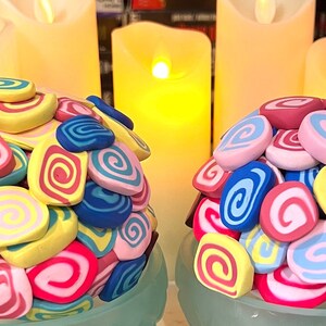 May include: Two decorative spheres made of colorful, swirled clay. Each sphere is composed of pink, blue, yellow, and red clay swirls. The spheres are sitting on a light blue surface.