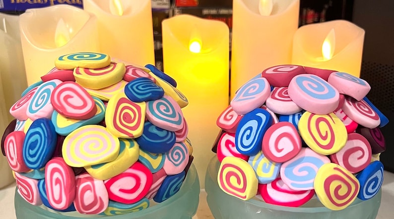 May include: Two decorative spheres made of colorful, swirled clay. Each sphere is composed of pink, blue, yellow, and red clay swirls. The spheres are sitting on a light blue surface.