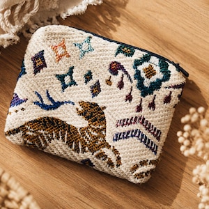 Op de afbeelding: Crèmekleurige, rechthoekige etui met ritssluiting en geborduurde details. Het ontwerp bevat een tijger, sterren en geometrische vormen in blauw, bruin en paars. De etui ligt op een houten oppervlak.