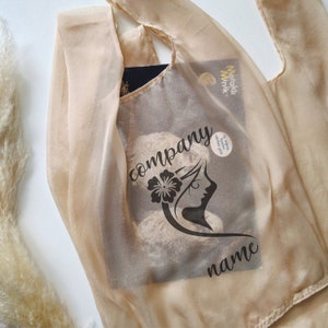 May include: A beige reusable tote bag with a black graphic design featuring a woman's face with a flower in her hair. The text "company name" is printed below the graphic. The bag is partially filled with a book.