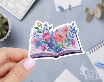 May include: A sticker featuring an open book illustration with a watercolor floral design. The flowers are pink, purple, and blue, with green leaves. The sticker has the text "BOMB DECAL" at the bottom.