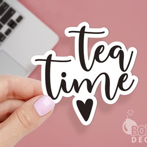K&ouml;nnte beinhalten: Wei&szlig;er Aufkleber mit schwarzem Text, der "tea time" lautet, mit einem kleinen schwarzen Herz unter dem Text. Der Aufkleber wird von einer Hand gehalten.