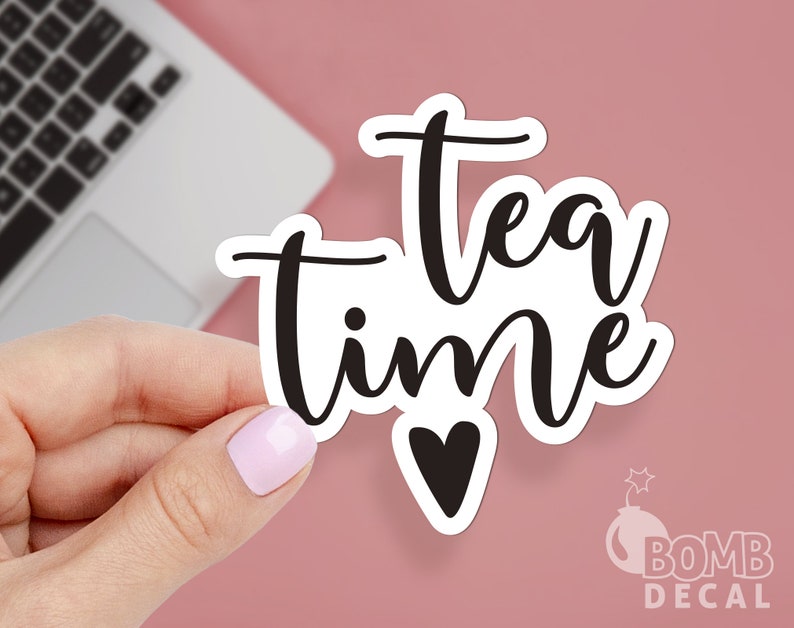 K&ouml;nnte beinhalten: Wei&szlig;er Aufkleber mit schwarzem Text, der "tea time" lautet, mit einem kleinen schwarzen Herz unter dem Text. Der Aufkleber wird von einer Hand gehalten.