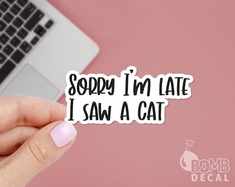 May include: White sticker with black text that reads "Sorry I'm late I saw a cat". The sticker is being held by a hand against a pink background.