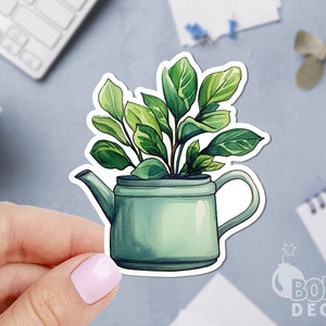 May include: A watercolor sticker featuring a green teapot filled with a leafy green plant. The sticker has a white border and is held by a hand with a light pink nail polish. The background is a light blue surface.