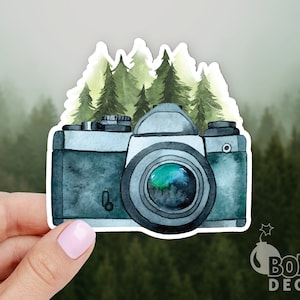 May include: A watercolor illustration of a vintage camera with a green forest background. The camera is gray and black with a large lens. The forest is a light green and yellow.