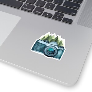 May include: A watercolor illustration of a vintage camera with a green and blue forest scene behind it. The camera is blue and black with a silver lens.