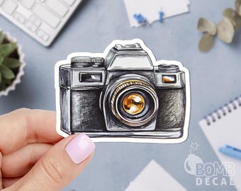 May include: A sticker featuring a detailed illustration of a vintage camera. The camera is rendered in shades of gray and black, with a prominent gold-colored lens. The sticker has a white border and the "BOMB DECAL" logo is visible.