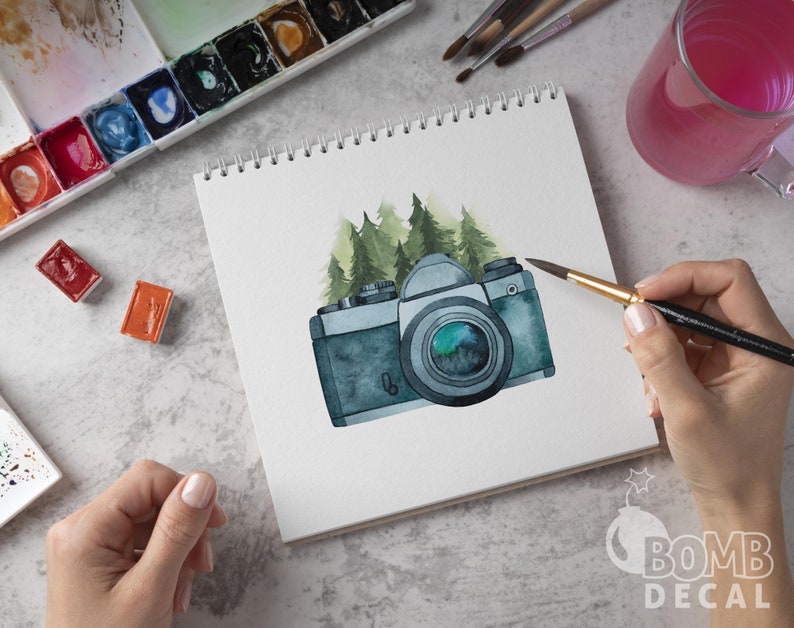 May include: A watercolor painting of a vintage camera with a green forest background. The camera is painted in shades of gray and blue, with a green lens. The forest is painted in shades of green and brown.