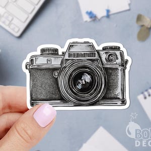 May include: A black and white sticker of a vintage camera. The camera has a large lens and detailed shading, giving it a realistic appearance. The sticker is held by a hand with a light pink nail polish.