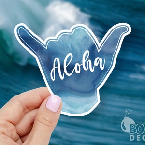 May include: A blue watercolor sticker with a hand making the shaka sign and the word "Aloha" written in white.