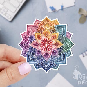 Könnte beinhalten: Ein farbenfroher Mandala-Aufkleber mit einem detaillierten floralen Design. Der Aufkleber zeigt ein Farbspektrum, darunter Rot, Orange, Gelb, Grün, Blau und Lila. Der Aufkleber wird von einer Person mit hellrosa Nagellack gehalten.