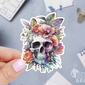 May include: A watercolor skull with a floral crown of pink, purple, and orange flowers. The skull is surrounded by green leaves and vines.