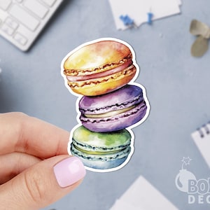 May include: A stack of three colorful macarons. The macarons are yellow, purple, and green. The macarons are arranged in a stack with the yellow macaron on top, the purple macaron in the middle, and the green macaron on the bottom.
