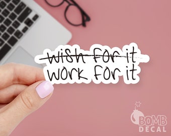 Op de afbeelding: Een witte sticker met zwarte tekst die "wish for it" zegt met een doorstreepte lijn en "work for it" eronder. De sticker wordt vastgehouden door een hand tegen een roze achtergrond.