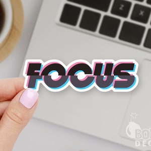 May include: A white sticker with the word "FOCUS" in black, pink, and blue. The sticker is held by a hand with a light pink nail polish, and is placed on a laptop.