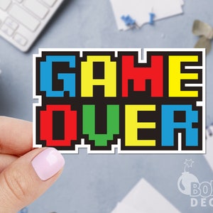 May include: A sticker with the words "GAME OVER" in pixelated, colorful letters. The letters are in blue, yellow, red, and green against a black background. The sticker is held by a hand.