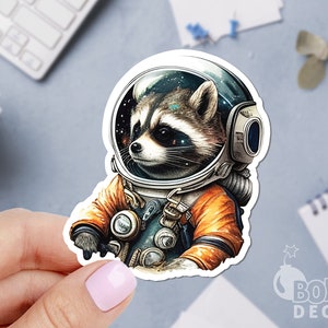 May include: A sticker featuring a raccoon in an astronaut helmet and spacesuit. The raccoon's face is visible inside the helmet, with a starry galaxy design. The spacesuit is orange and teal. The sticker has the text "BOMB DECAL".