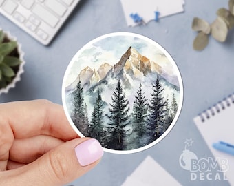 May include: A round sticker featuring a watercolor painting of a mountain range with pine trees in the foreground. The mountain peaks are brown and tan, with a misty, blue-gray sky. The sticker has the text "BOMB DECAL".