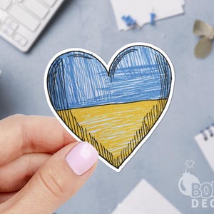 Könnte beinhalten: Ein herzförmiger Aufkleber mit einer handgezeichneten Illustration, die einen blauen oberen und einen gelben unteren Bereich aufweist, welche die Farben der ukrainischen Flagge mit sichtbaren Stiftstrichen und einer schwarzen Umrandung nachahmen.
