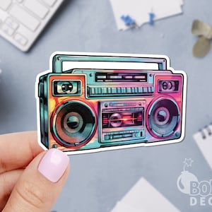May include: A colorful sticker featuring a retro boombox design. The boombox has a vibrant color scheme with shades of pink, blue, orange, and green. The sticker has a white border and is held by a hand.