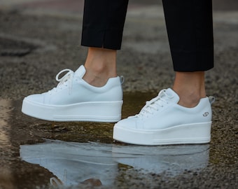 May include: White leather platform sneakers with a lace-up closure. The sneakers have a thick white sole and are shown being worn by a person.