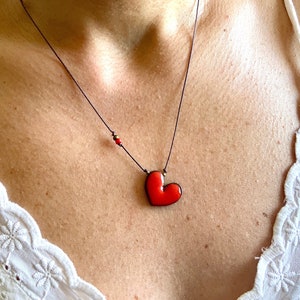 Puede incluir: Un collar con un colgante en forma de corazón rojo en un cordón negro. El colgante está hecho de cerámica y tiene un acabado brillante.