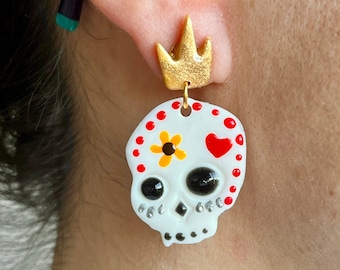 May include: White sugar skull earrings with red and yellow details, a small red heart, and a gold crown charm.