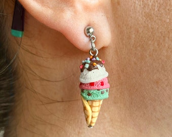 Puede incluir: Un par de pendientes colgantes con un cono de helado en miniatura con tres bolas. El helado está en capas de verde, rosa y blanco, con chispas de chocolate y un chorrito de chocolate en la parte superior. El cono es un cono de waffle marrón claro.
